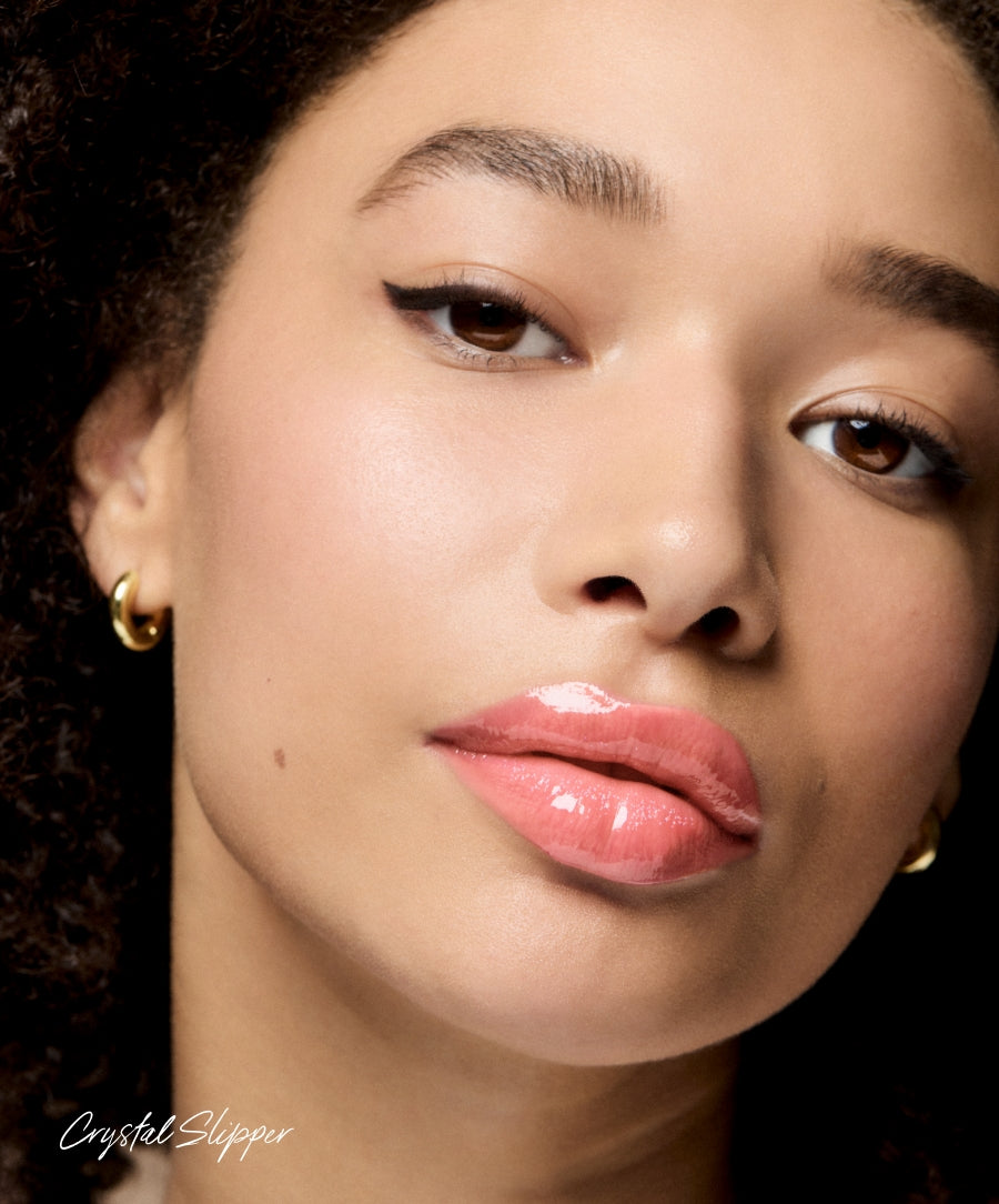 Close-up of a woman wearing shiny pink glossy Legendary Lip Oil with smooth, natural makeup and gold hoop earrings, highlighting clean beauty and skincare infused makeup.
