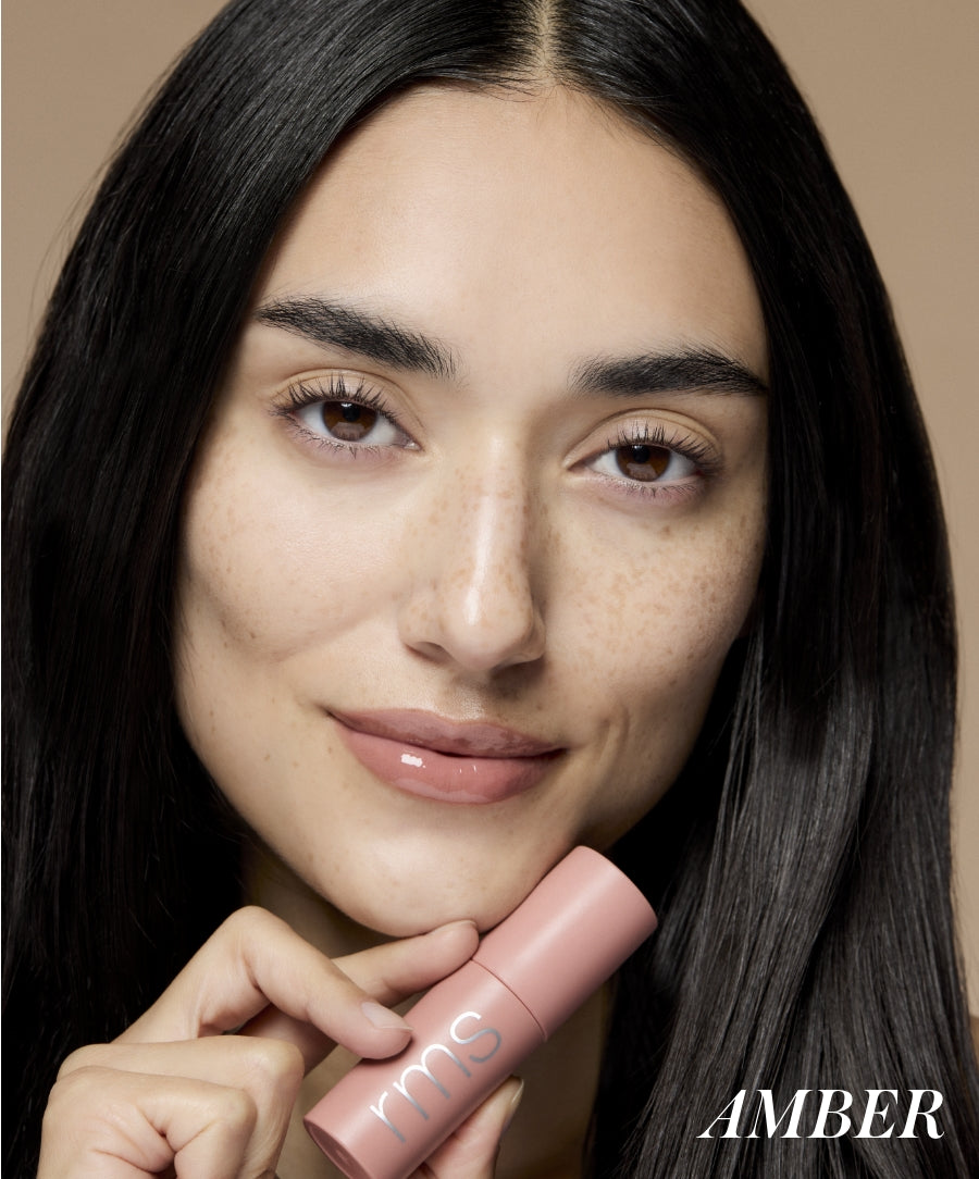 Close-up of a woman with clear skin and natural makeup holding a pink tube of Legendary Lip Oil, showcasing glossy lips and a fresh, radiant look.