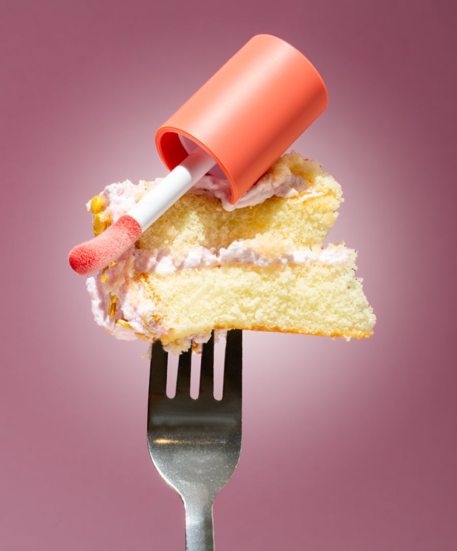 A glossy applicator with coral-colored Legendary Lip Oil resting on a fork holding a slice of yellow cake with light pink frosting against a soft pink background.