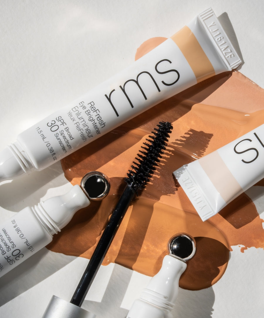 Close-up of RMS Beauty makeup products including an eye brightener with applicator and mascara wand, displayed on a neutral background with warm beige liquid makeup swatches, featuring The Wide Awake Duo.
