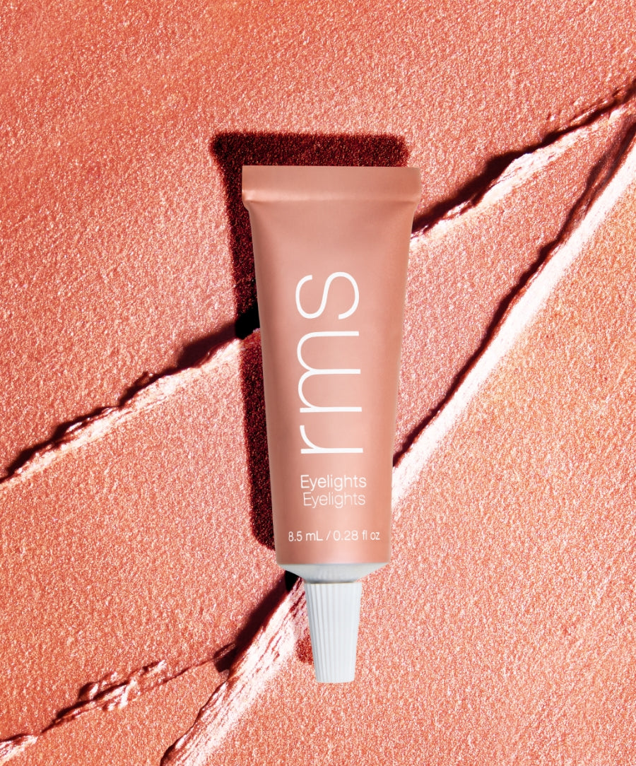 Tube of Eyelights Cream Eyeshadow in a warm peachy pink shade on a textured, softly shimmering background, highlighting clean beauty and natural ingredients.