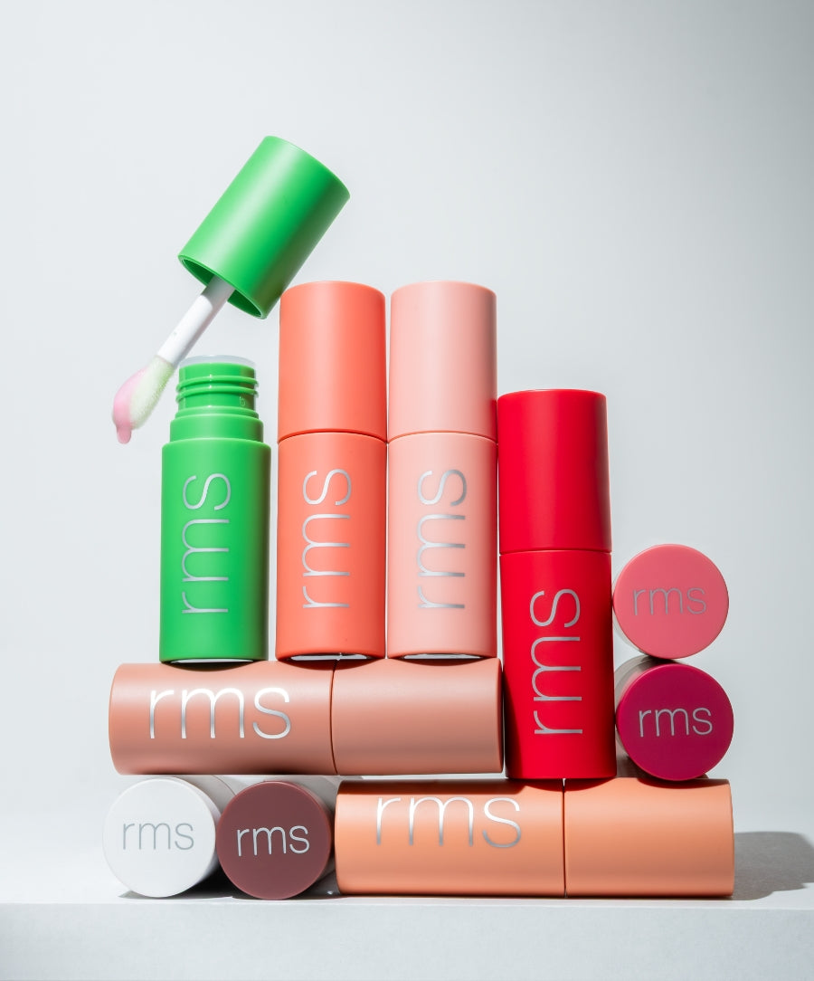 Collection of colorful rms lip balm tubes arranged in a stack with one green tube open showing the applicator, featuring the Legendary Lip Oil.