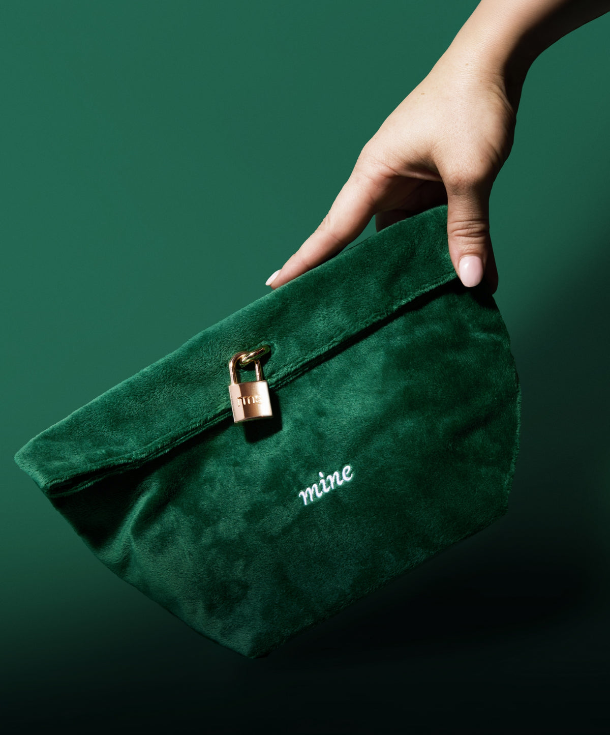 Hand holding a dark green velvet pouch with a small gold lock and the word "mine" embroidered, part of The Chameleon 2-Piece Set, emphasizing clean beauty and sustainable packaging.