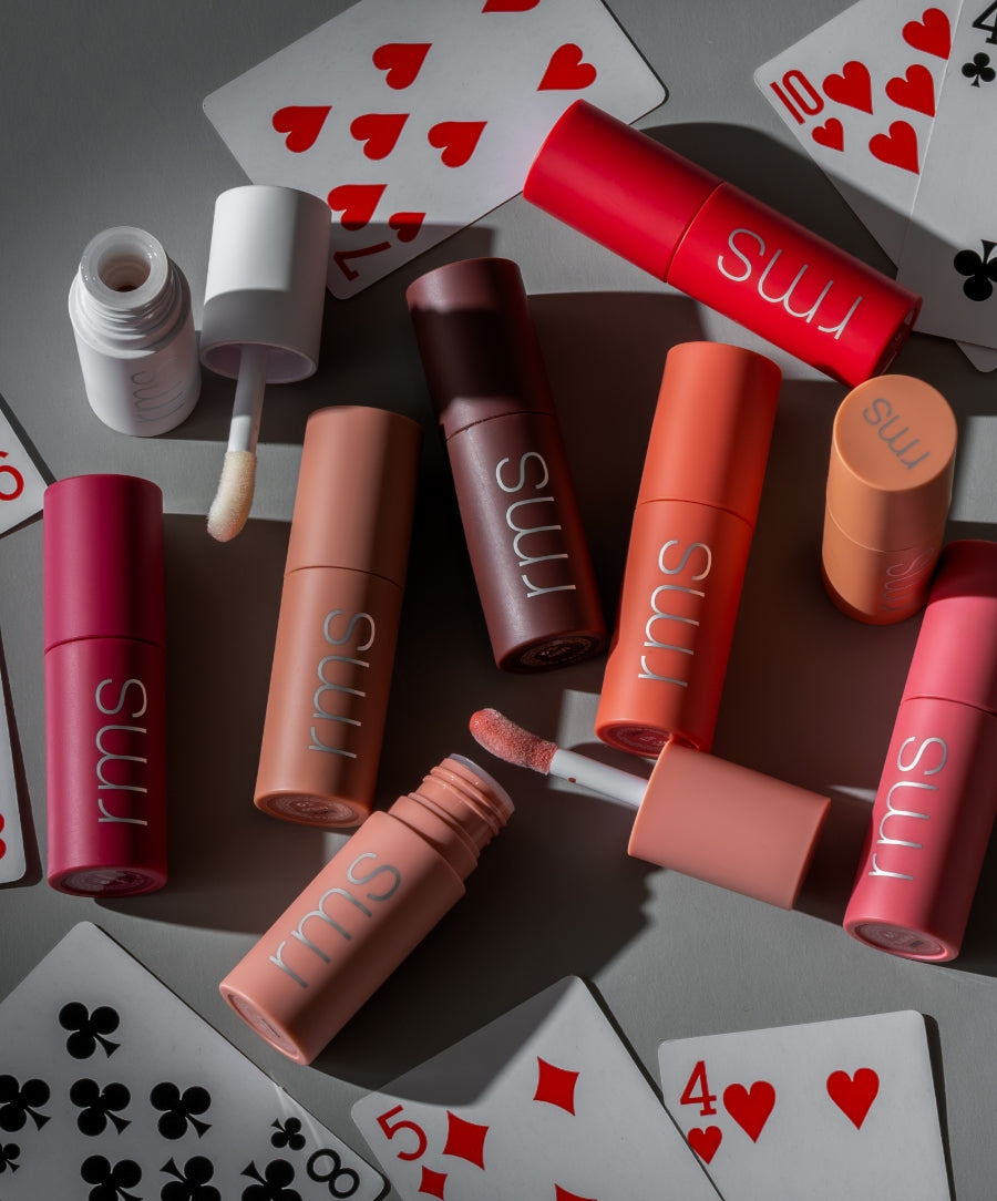 Array of RMS Beauty Legendary Lip Oil tubes in various pink, peach, and red shades surrounded by playing cards, highlighting clean beauty and natural ingredients.