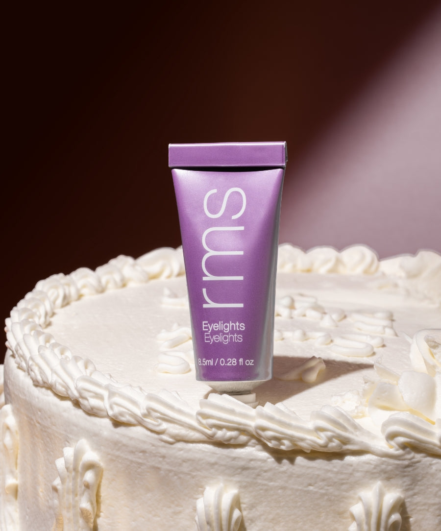 A tube of Eyelights Cream Eyeshadow in a soft purple shade standing upright on a white frosted cake with decorative icing, highlighting clean beauty and natural ingredients.