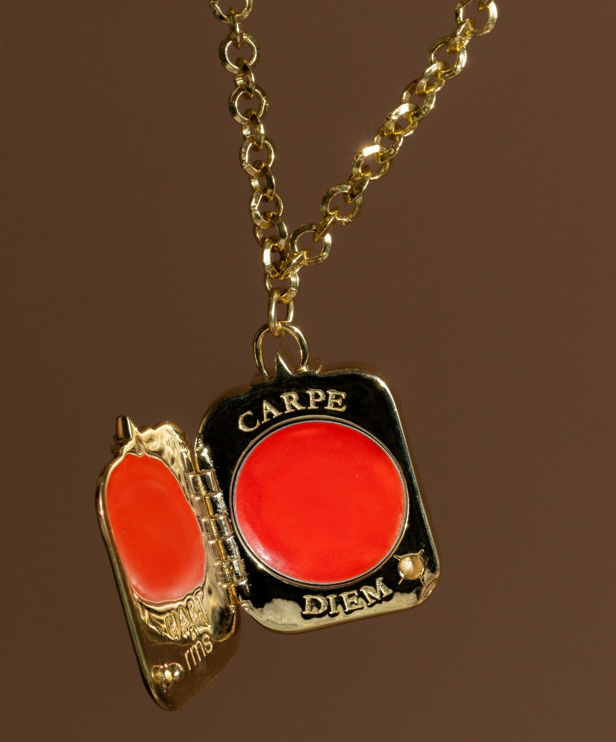 HART x RMS: Lip2Cheek Locket featuring a gold chain with a small, square-shaped locket containing vibrant coral lip and cheek color, engraved with "CARPE DIEM" on the front.