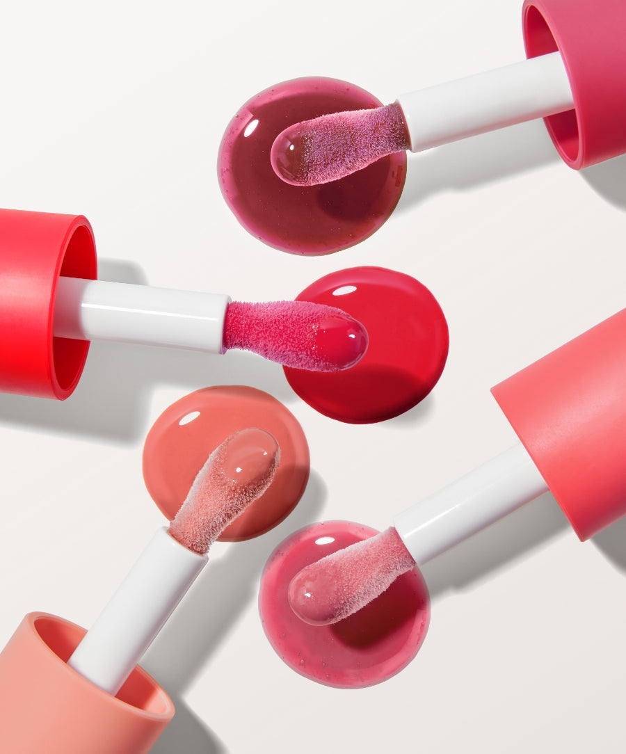 Four applicators with different shades of Legendary Lip Oil resting on matching color pools on a white background, showcasing the product’s glossy texture and natural ingredients.