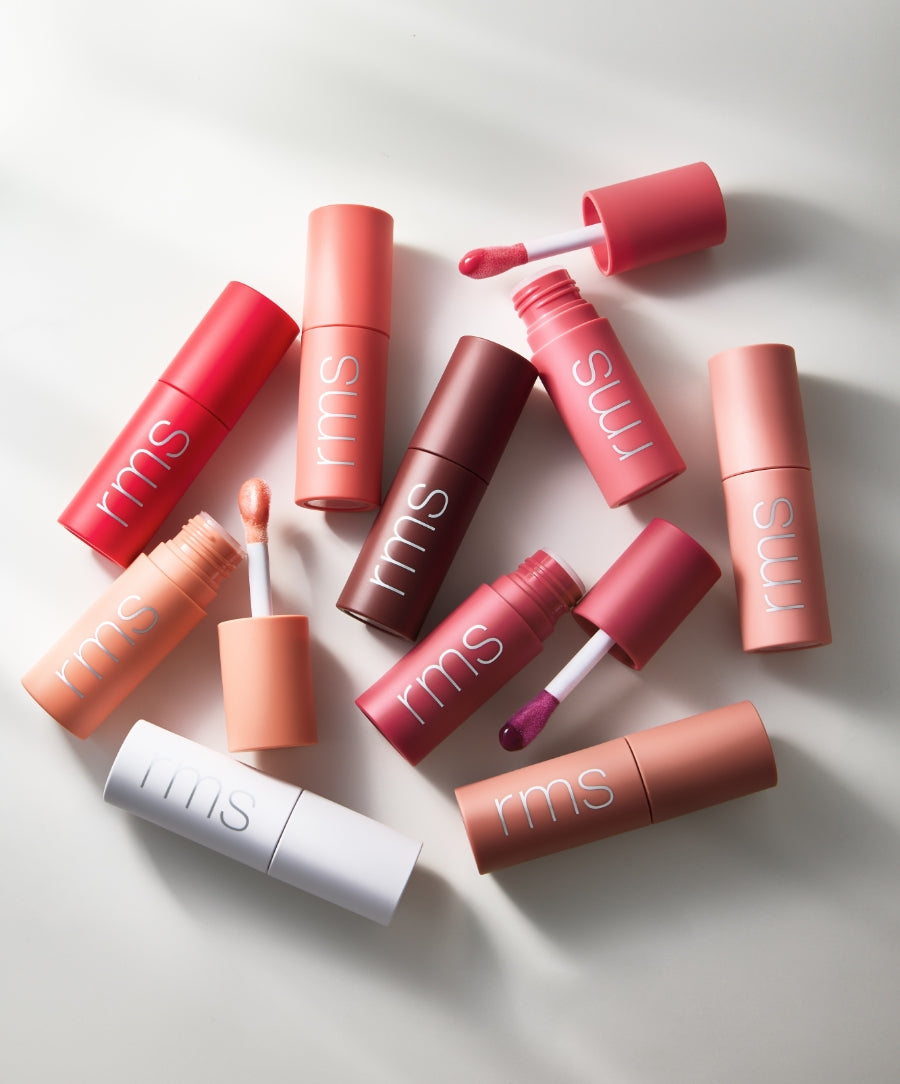 Collection of colorful RMS Beauty tubes of Legendary Lip Oil with some open showing glossy applicators in natural light on a white surface, highlighting clean beauty and natural ingredients.