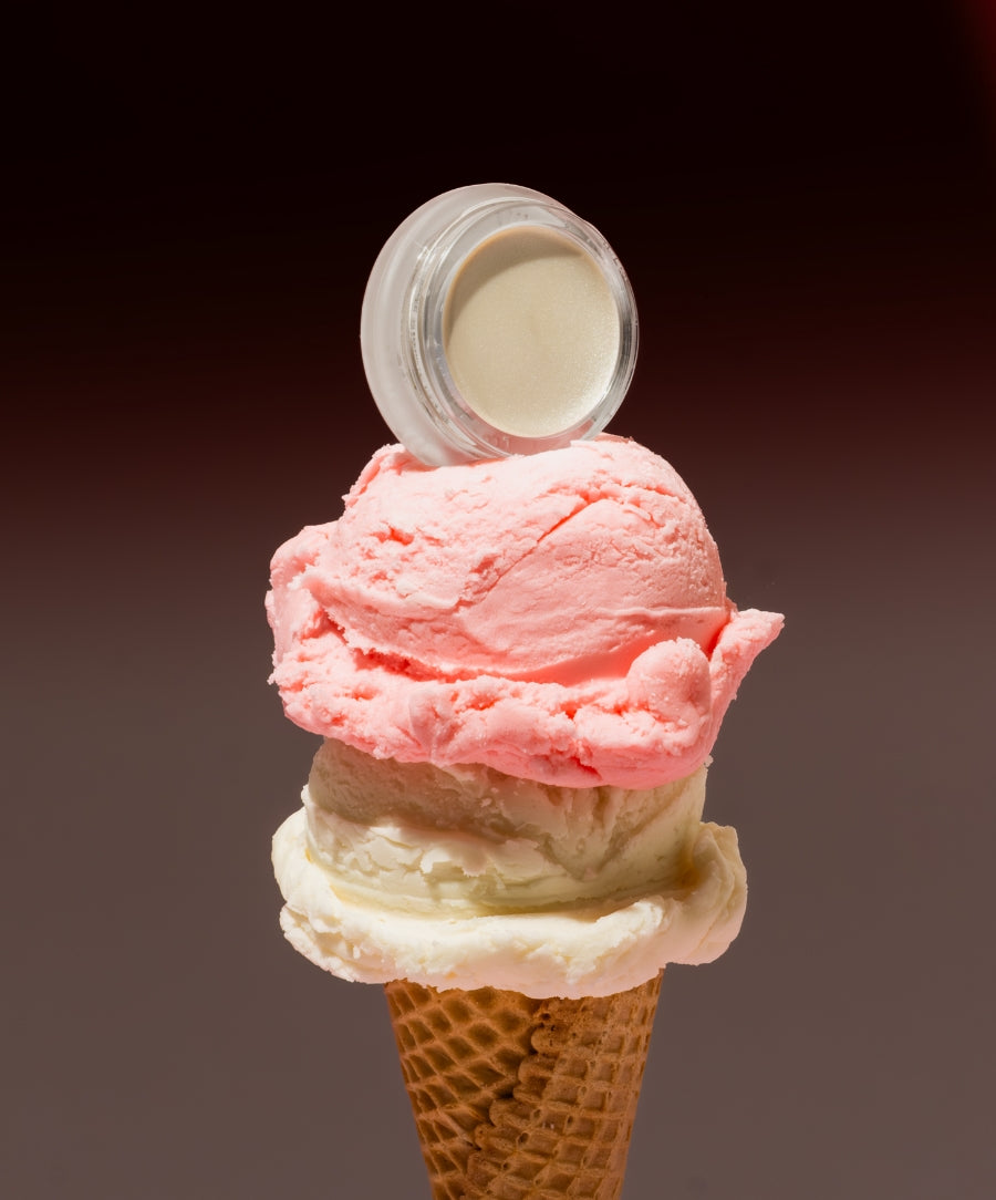 Luminizer cream makeup jar balanced on top of a double scoop ice cream cone with pink and white scoops against a gradient dark background, evoking fresh and natural beauty.