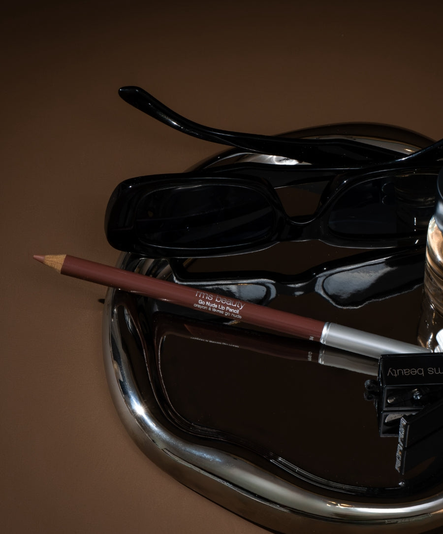 Go Nude Lip Pencil displayed on a reflective surface next to black sunglasses and a pencil sharpener, highlighting clean beauty and sustainable packaging.