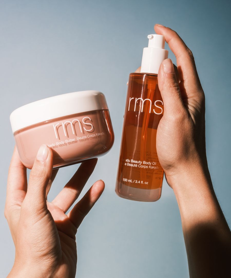 Hands holding RMS Beauty skincare products, featuring a creamy body butter and a golden body oil, highlighting The Radiant Body Duo set against a soft blue background.