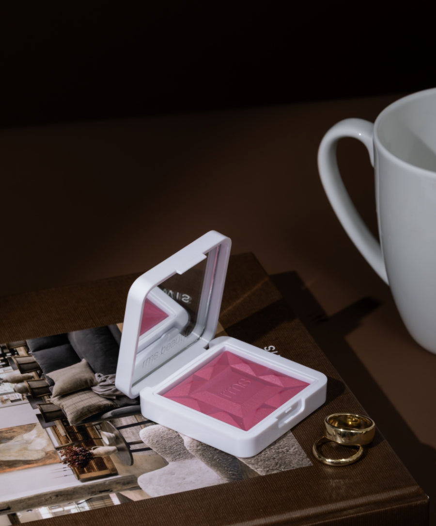 Compact white case with a pink ReDimension Hydra Powder Blush featuring a geometric pattern, placed on a book next to gold rings and a white ceramic cup, showcasing clean beauty makeup.