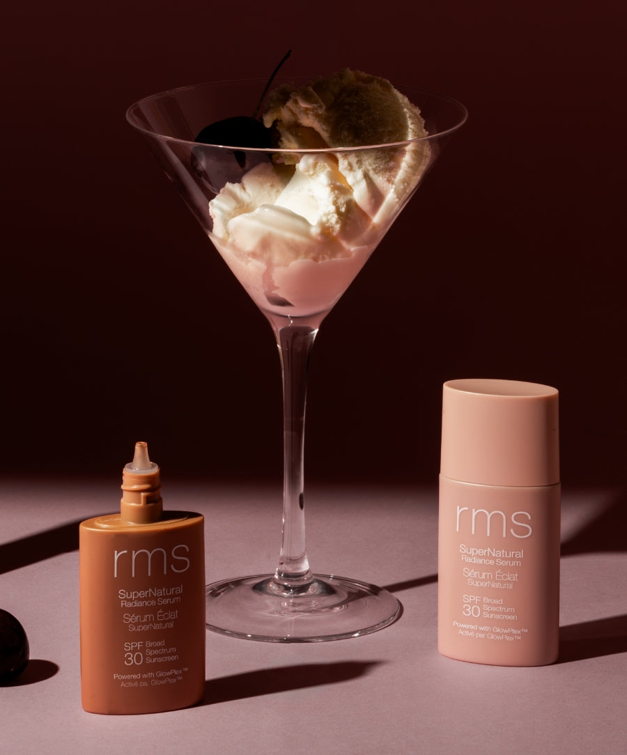 Two bottles of Supernatural Radiance Tinted Serum with SPF 30 by RMS Beauty displayed next to a dessert glass with whipped cream and a cherry, highlighting clean beauty and natural ingredients.