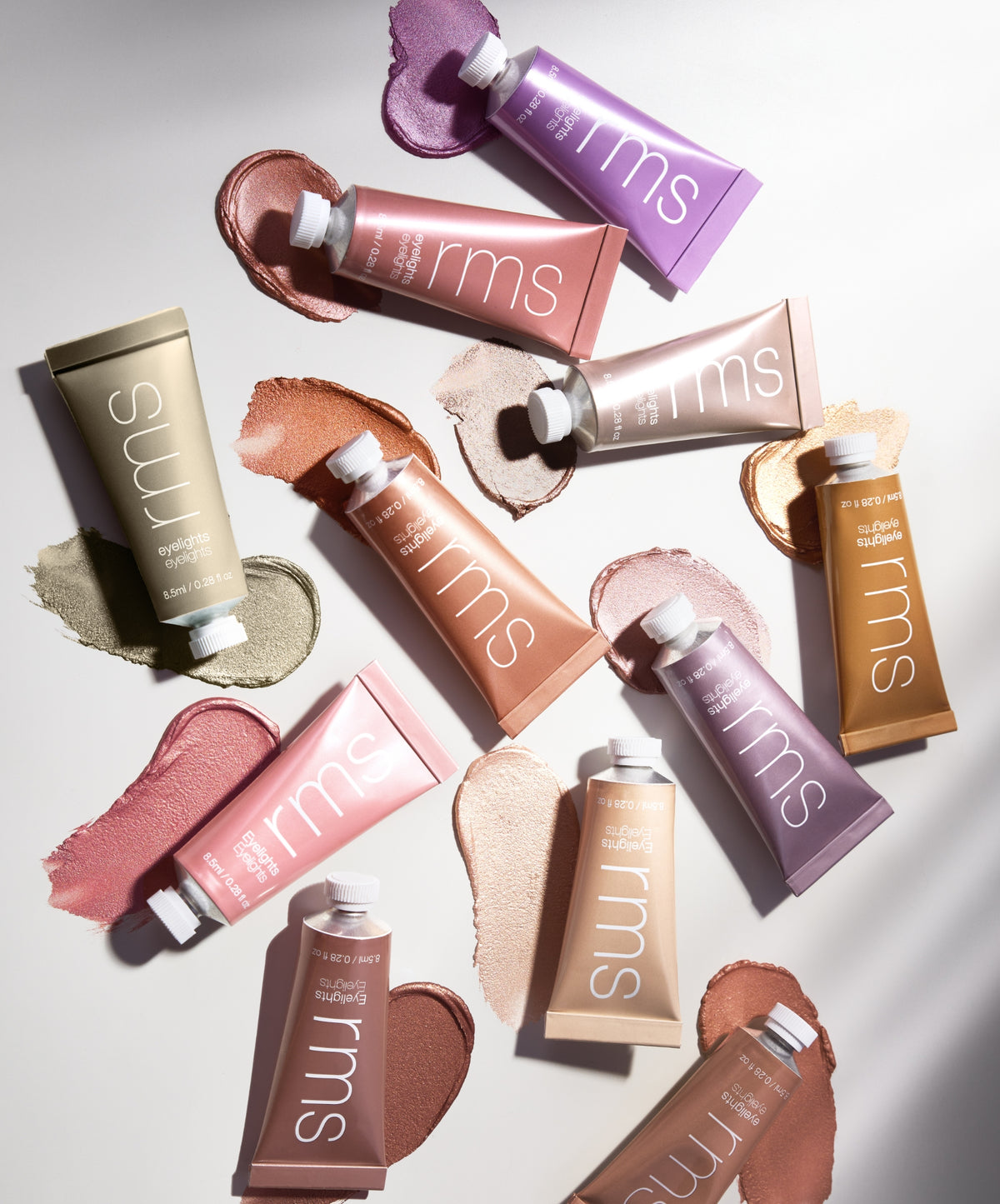Various colorful tubes of Eyelights Cream Eyeshadow from RMS Beauty arranged with swatches of different shades on a white surface, showcasing creamy texture and clean beauty makeup.
