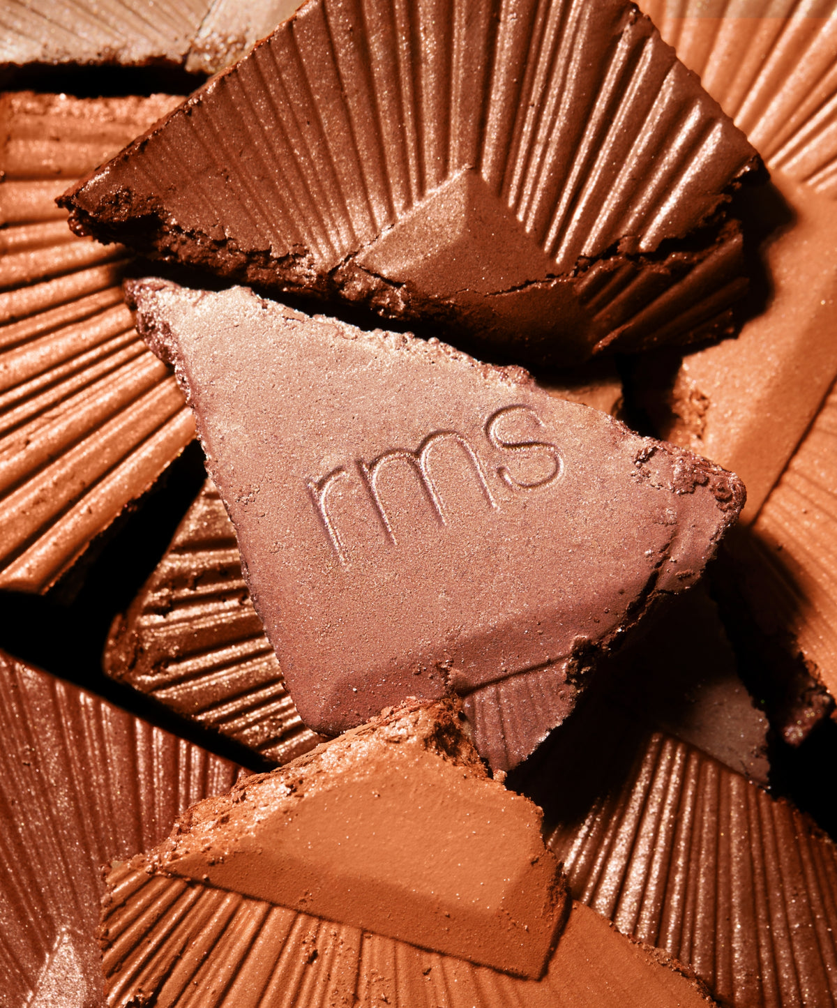 Close-up of various shades of broken pressed powder bronzer pieces, featuring one with "rms" embossed, highlighting the ReDimension Hydra Bronzer texture and natural ingredients.
