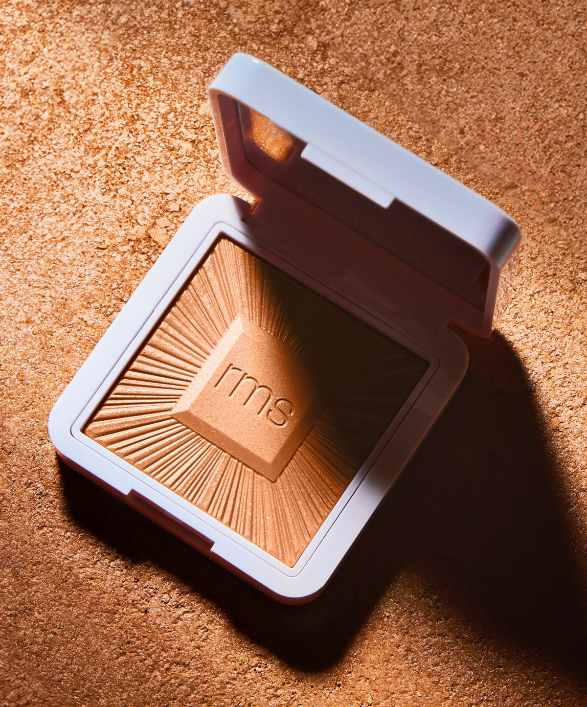 Open compact of ReDimension Hydra Bronzer by RMS Beauty with a sunburst embossed design on a shimmering bronze background, highlighting clean beauty and natural ingredients.