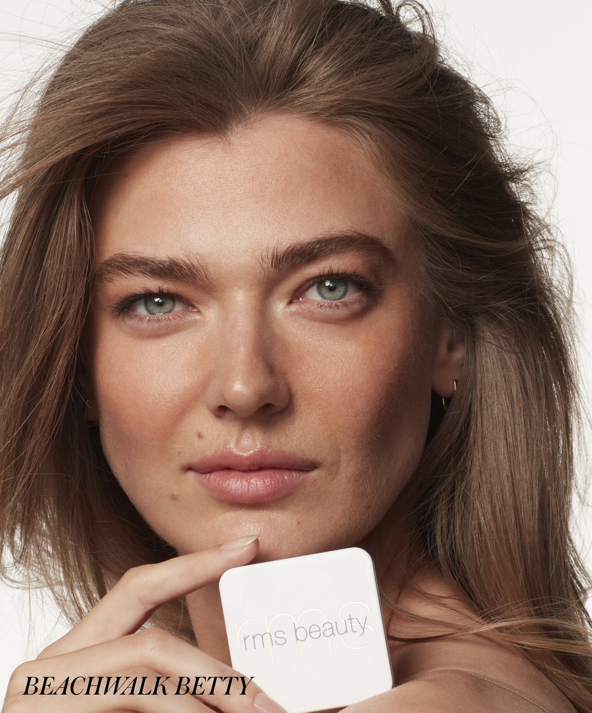 Close-up of a woman with natural makeup holding a white compact of ReDimension Hydra Bronzer by RMS Beauty, highlighting clean beauty and skincare-infused makeup.
