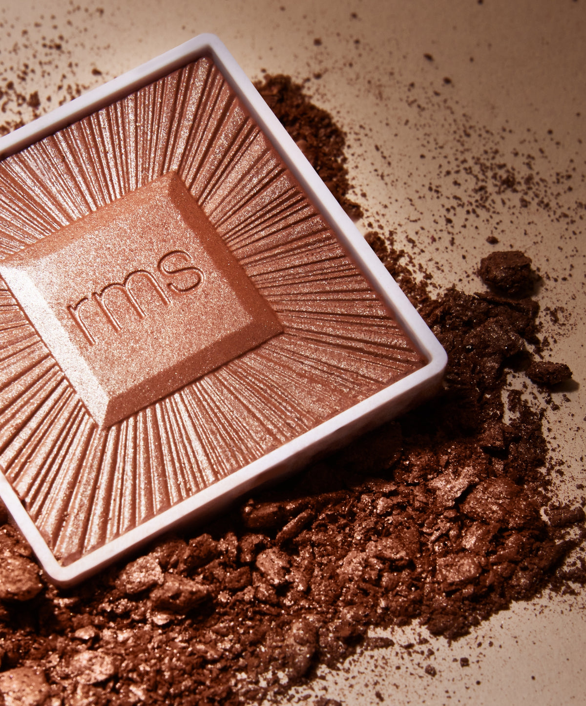 Close-up view of a shimmering bronzer with "rms" embossed on its surface, surrounded by crushed bronzer powder in warm, natural tones, showcasing the ReDimension Hydra Bronzer.