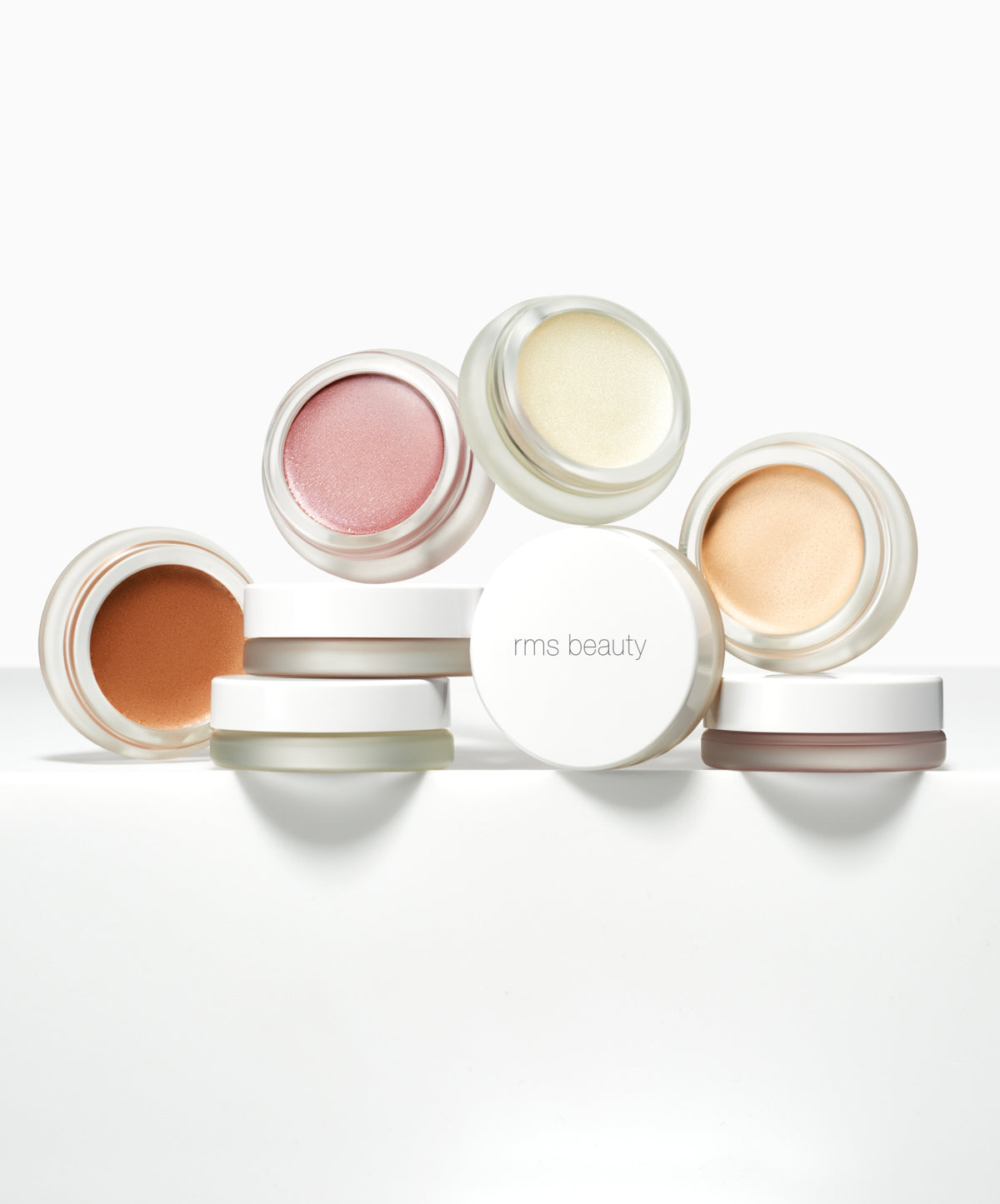 Collection of small open jars containing various shades of Luminizer cream makeup with a white jar lid featuring a minimalist design on a clean white background