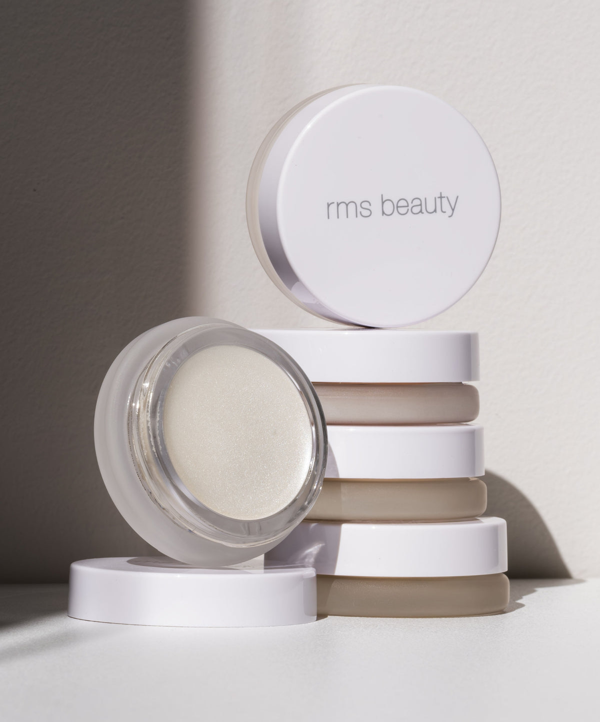 Stack of white RMS Beauty containers with one open jar showing a shimmering cream product, highlighting the Luminizer with clean beauty and natural ingredients.