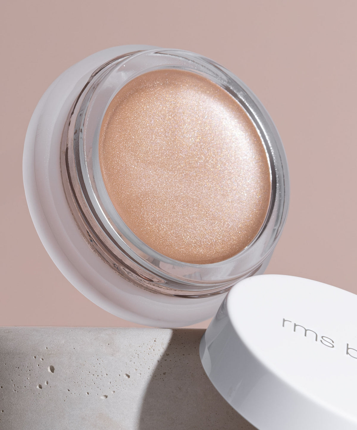 Close-up of Luminizer cream highlighter in a transparent glass jar with a white lid resting on a neutral-toned surface, showcasing its smooth, shimmering champagne shade.