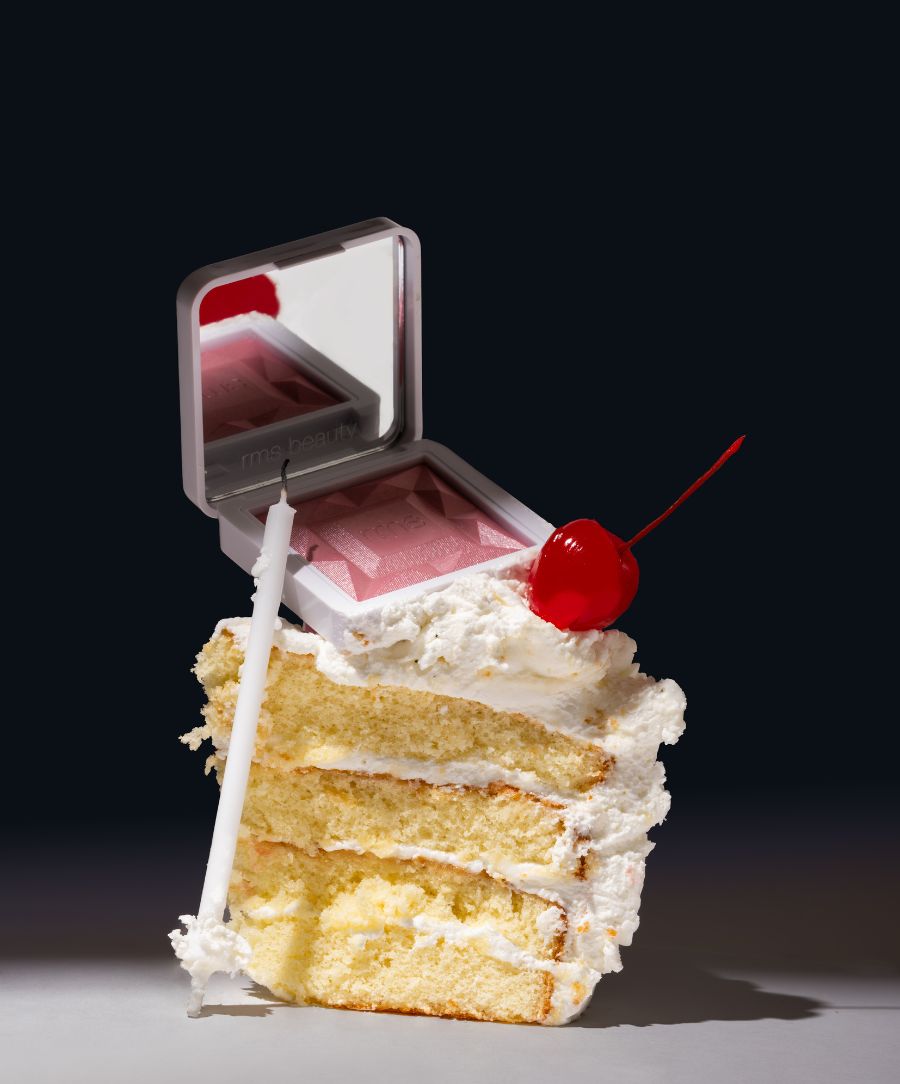 Slice of vanilla layer cake with white frosting and a cherry, topped with an open compact of ReDimension Hydra Powder Blush and a white birthday candle.
