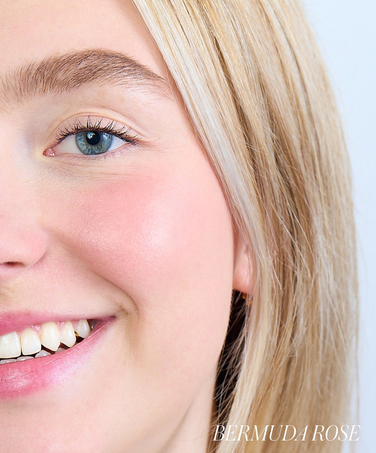 Close-up of a smiling woman with fair skin and blonde hair wearing ReDimension Hydra Powder Blush in a natural rosy shade on her cheeks, showcasing a clean beauty makeup look.
