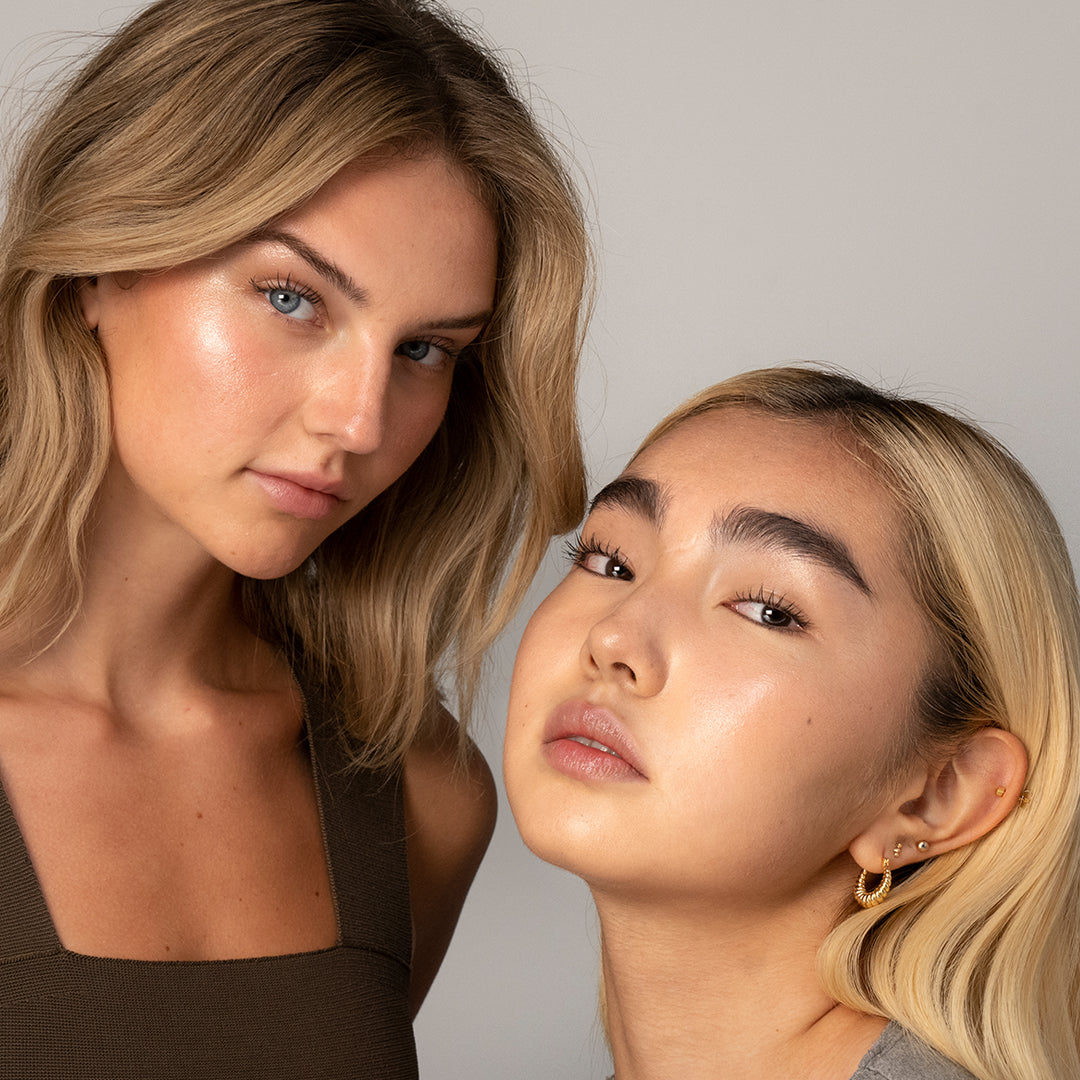Two women with glowing, radiant skin showcasing the luminous effect of Luminizer in a natural makeup look against a neutral background.
