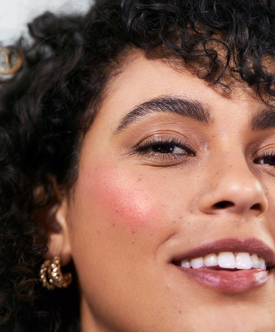 Close-up of a smiling woman with natural curly hair wearing ReDimension Hydra Powder Blush on her cheeks, showcasing a radiant, clean beauty look with natural ingredients.