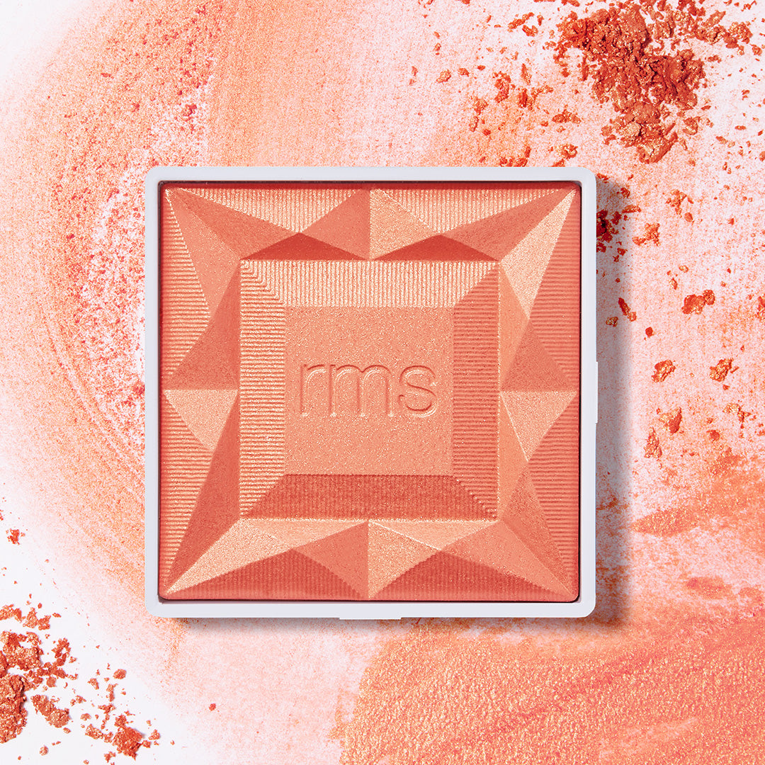 Square compact with geometric embossed design in warm coral shades and the embossed initials rms, surrounded by crushed powder highlighting the ReDimension Hydra Powder Blush from RMS Beauty, featuring clean ingredients and sustainable packaging.