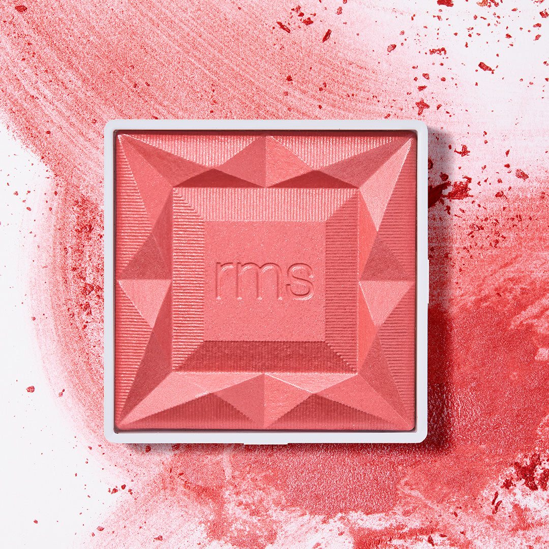 ReDimension Hydra Powder Blush in a vibrant coral shade, featuring a geometric embossed design with RMS Beauty branding, surrounded by scattered powder for a clean beauty makeup look.